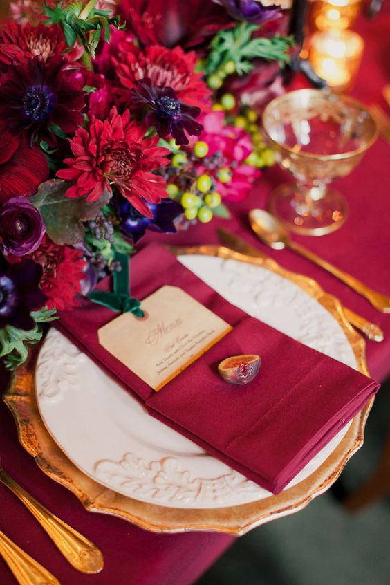 a colorful fall tablescape with marsala linens, bold red and marsala blooms, gold cutlery and gold rimmed glasses