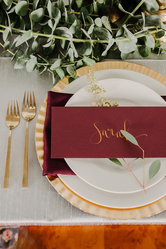 a fresh wedding tablescape with a lush greenery runner, gold cutlery and a charger, a marsala napkin and a card