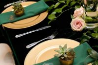 a gold and emerald wedding tablescape with a black tablecloth, gold chargers, emerald napkins, potted succulents and pink roses and greenery