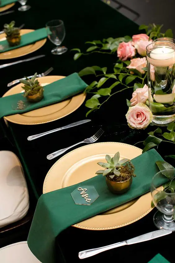 a gold and emerald wedding tablescape with a black tablecloth, gold chargers, emerald napkins, potted succulents and pink roses and greenery