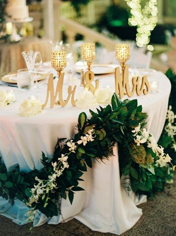 a lovely wedding sweetheart table with a lush greenery and white bloom garland, gold calligraphy and candleholders and white porcelain