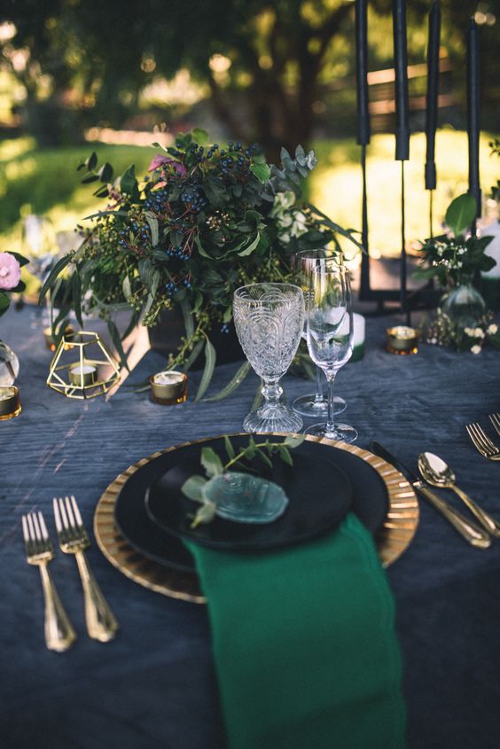 a moody wedding tablescape with a graphite grey tablecloth, gold placemats and cutlery, a greenery and berry centerpiece, gold candleholders