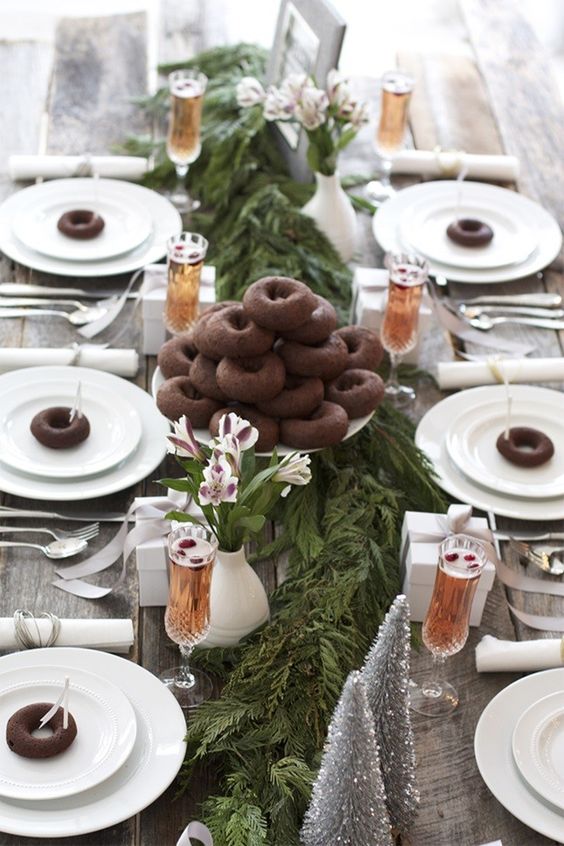 a natural tablescape with a fresh greenery runner, silver trees, chocolate donuts to make each place setting