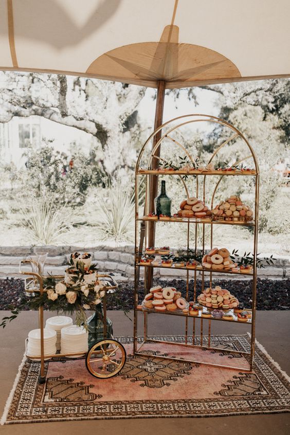 a pretty brass shelving unit with lots of sweets and greenery decor, a small trolley with a wedding cake is amazing