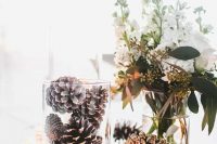 a romantic winter wedding centerpiece of candles, pinecones in glasses and white blooms and greenery in a vase