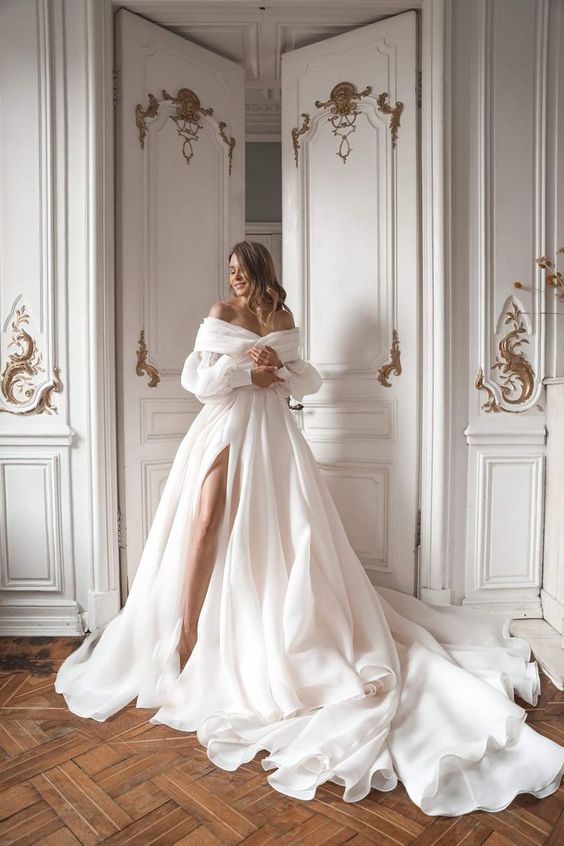 a simple yet elegant princess wedding ballgon with an off the shoulder neckline and puff sleeves, a pleated and layered skirt with a train and a thigh high slit