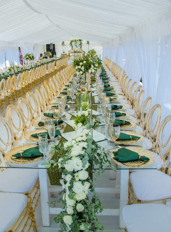 a sophisticated emerald and gold wedding tablescape with a greenery and white runner, emerald napkins, gold placemats and gold-rimmed glasses