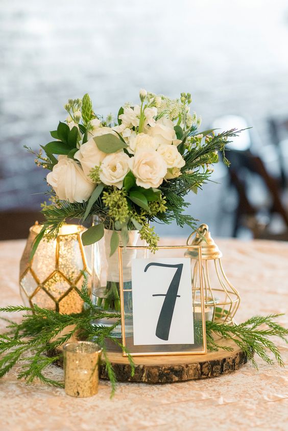 a stylish emerald and gold wedding centerpiece of a wood slice, gold candleholders and a lantern, neutral blooms with greenery