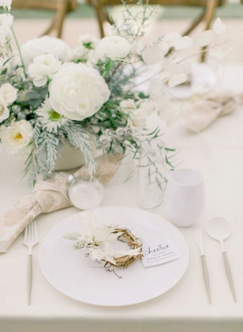 a stylish frozen tablescape with white blooms and greenery, mini wreaths with feathers and white porcelain and cutlery