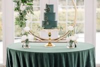 a stylish wedding cake table with an emerald tablecloth, a blue cake with leaves, a gold arch with greenery and blooms