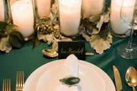 a stylish wedding table setting with an emerald tablecloth, gold cutlery, greenery and candles on the table