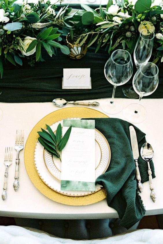 a stylish wedding tablescape with emerald linens, greenery and neutral blooms, gold placemats, green menus and greenery