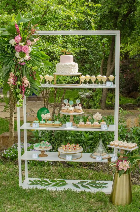 a tall shelving unit decorated with pink tropical blooms and fronds and with lots of delicious desserts on display is an amazing idea