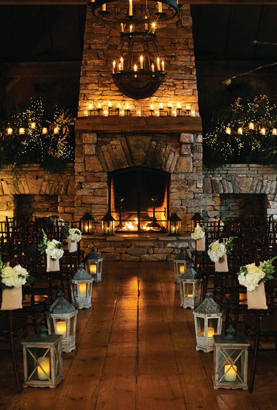a white flower arrangements with greenery, candle lanterns to decorate the wedding aisle and a cozy fireplace with lots of candles as a backdrop