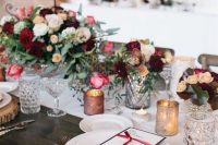 a winter wedding tablescape with a white runner, bold neutral and marsala blooms, greenery, wood slices and marsala napkins plus candles