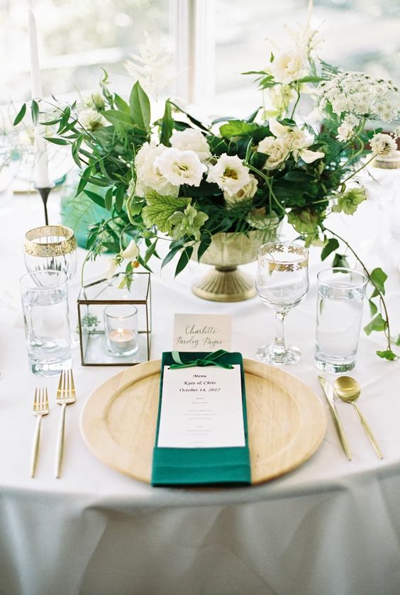 an airy wedding tablescape with a neutral tablecloth, wooden placemats, green napkins, a greenery and white bloom centerpiece and gold rimmed glasses