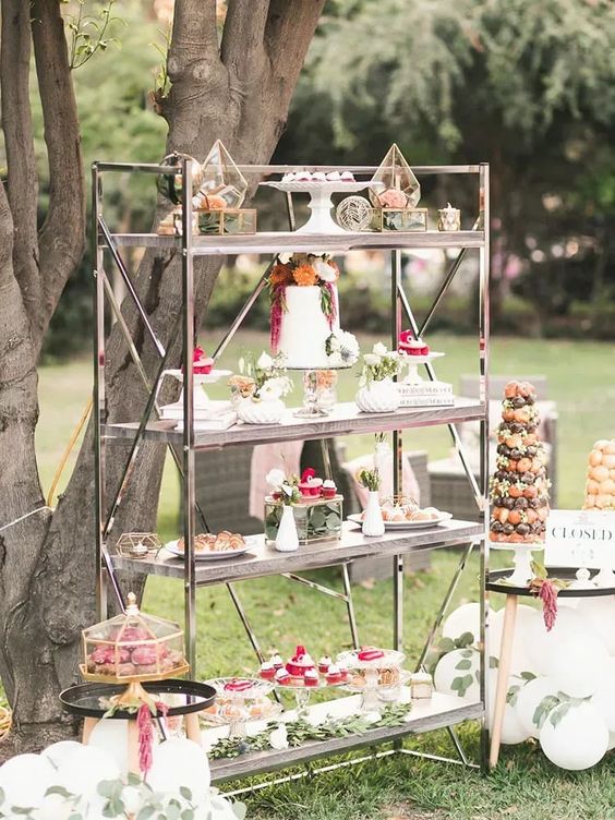 an elegant vintage shelving unit with lots of desserts, grenery and bright blooms is a stylish idea for a garden wedding