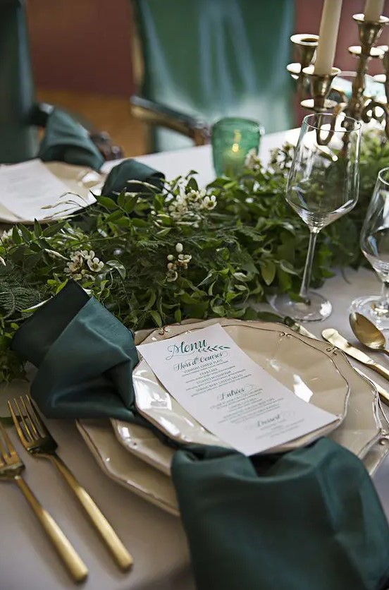 emerald napkins and candleholders, gold cutlery and a lush greenery garland in the center