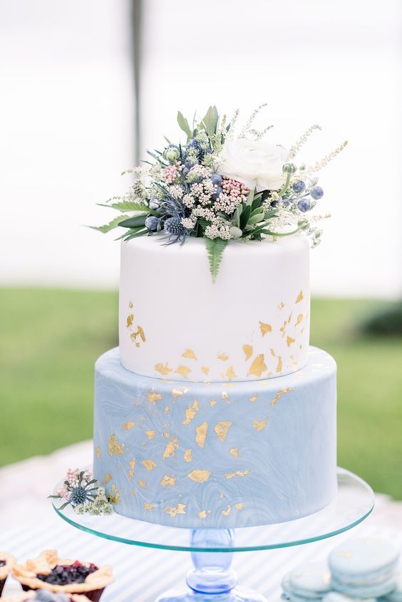 a blue marble and white wedding cake with gold foil and white and blue blooms and berries on top plus some foliage