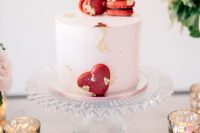 a blush wedding cake decorated with gold leaf and red heart-shaped macarons is a cool idea for a Valentine wedding