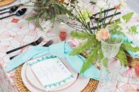 a bold tropical wedding tablescape with a printed tablecloth, a woven charger, greenery and blooms, candles and black cutlery