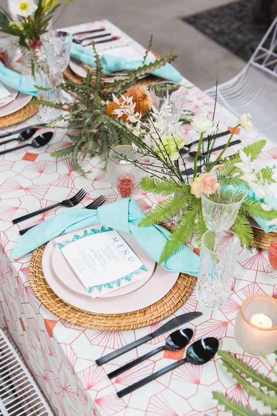 a bold tropical wedding tablescape with a printed tablecloth, a woven charger, greenery and blooms, candles and black cutlery
