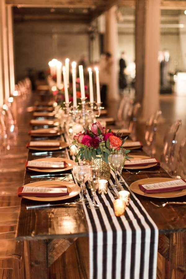 a bold wedding tablescape with a striped table runner, bold blooms, candles, gold chargers and burgundy napkins