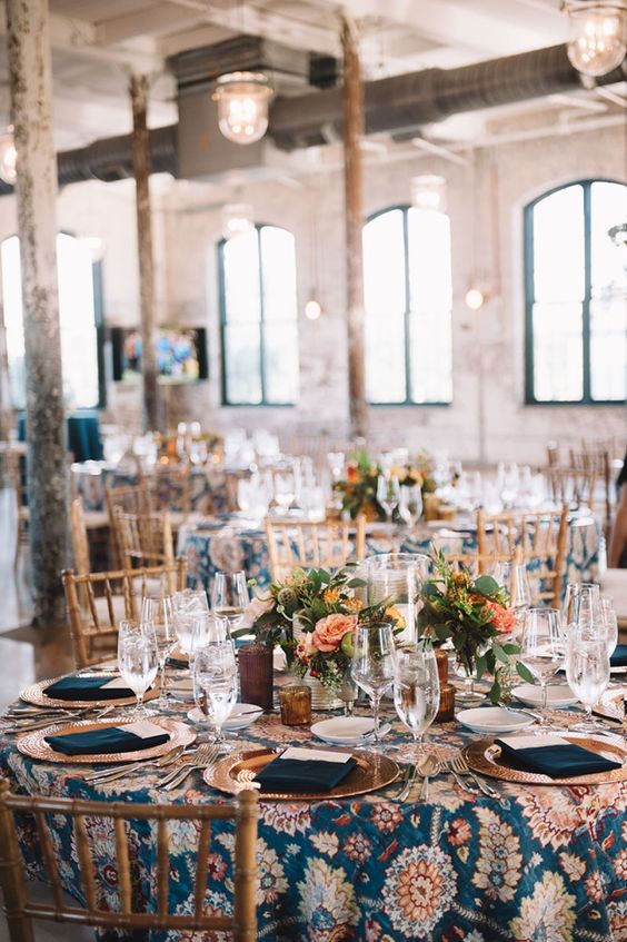 a bright and whimsical wedding tablescape with a printed tablecloth, orange blooms and greenery, navy napkins and copper chargers