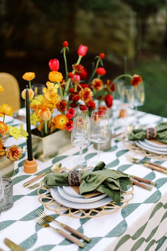 a bright wedding tablescape with a bold printed tablecloth and green napkins, bold blooms in planters, black candles, gold cutlery and candleholders