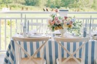 a coastal wedding tablescape with a blue printed tablecloth, a refined floral centerpiece and a decorated piece of driftwood over the table