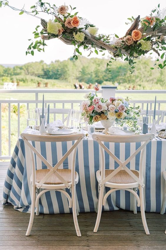 a coastal wedding tablescape with a blue printed tablecloth, a refined floral centerpiece and a decorated piece of driftwood over the table