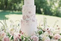 a delicate spring wedding cake with pink and mauve blooms painted and lush blooms around is amazing