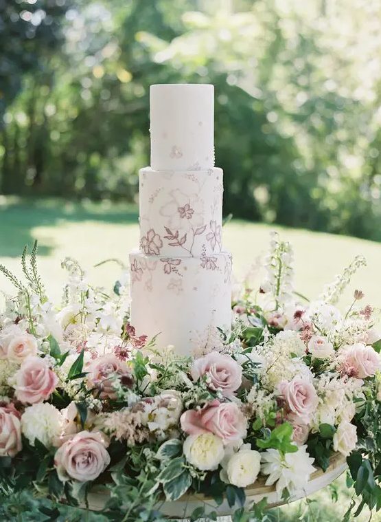 a delicate spring wedding cake with pink and mauve blooms painted and lush blooms around is amazing