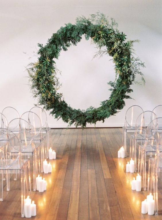 a minimalist wedding ceremony space with a giant greenery wedding wreath, sheer chairs and candles lining up the aisle