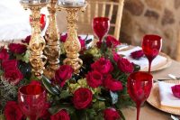 a refined Valentine’s Day wedding centerpiece of greenery, red roses, refined gold candleholders and red glasses to match