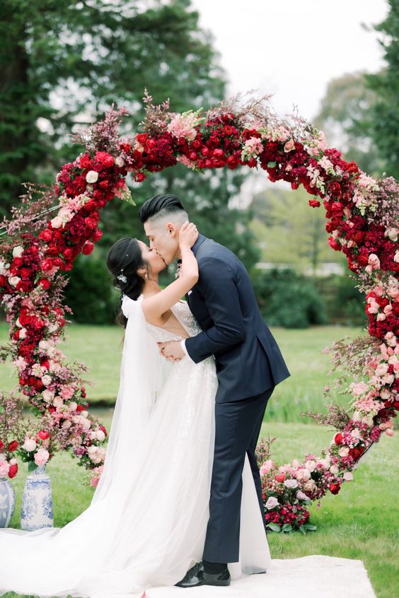 a round wedding arch covered with red roses, pink and blush blooms and some blooming branches for a Valentine wedding