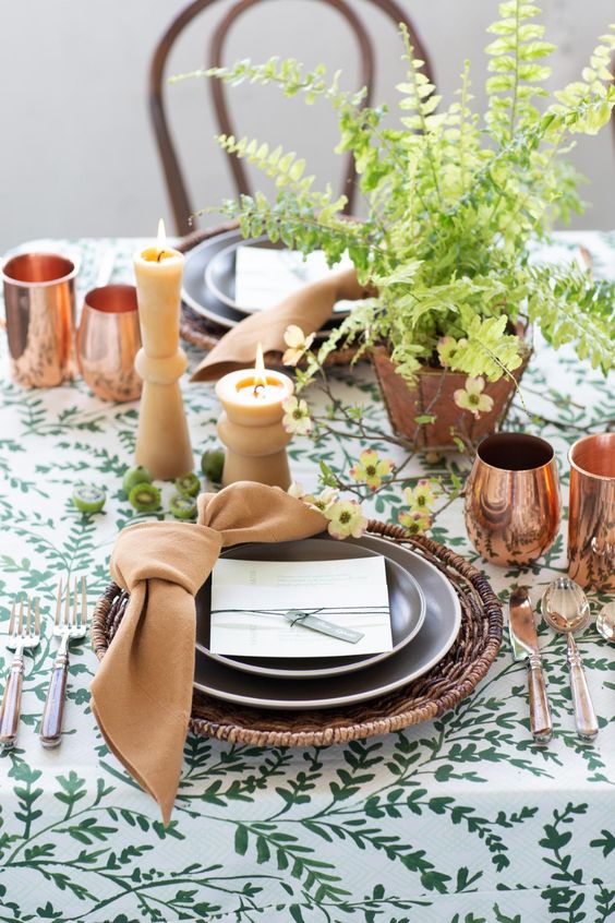 a spring wedding tablescape with a printed tablecloth, potted greenery, a tan naokin and candles, copper mugs