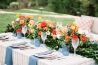 a summer wedding tablescape with a plaid tablecloth and blue napkins, a greenery and bright flower table runner, blue glasses