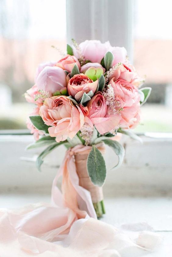 a sweet pastel Valentine wedding bouquet pf peachy and light pink blooms and greenery and light pink ribbons