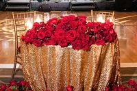 a sweetheart table with a gold sequin tablecloth and lots of red roses and greenery on the table and on the floor