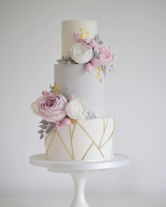 a white and dove grey wedding cake with gold geometric patterns, white and blush blooms, gold foil and pale greenery