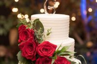 a white buttercream wedding cake topped with red roses, greenery and a silver monogram is a lovely dessert