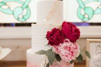 a white to pink ombre wedding cake decorated with gold leaf, with burgundy and pink blooms and greenery