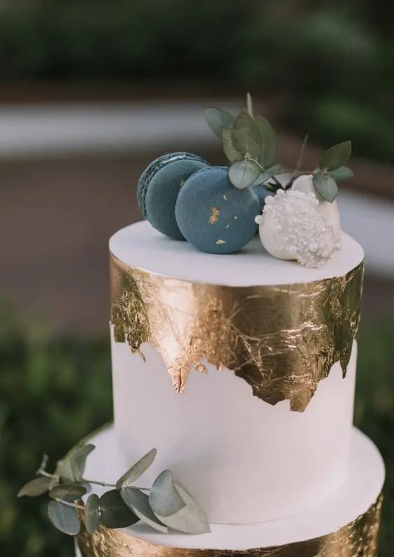 a white wedding cake with gold leaf, blue and white macarons and eucalyptus looks very edgy