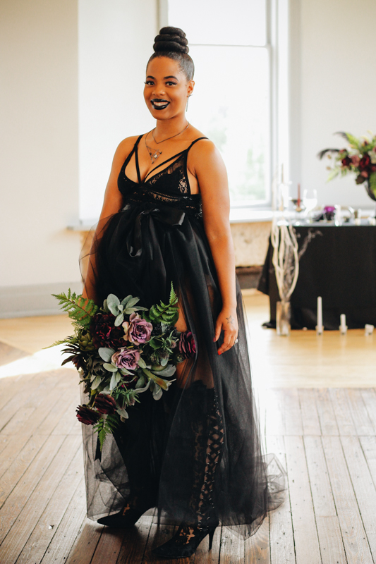 a Halloween bride wearing an A-line black wedding dress with a lace bodice with straps, a sheer skirt and tall lace boots