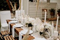 a beautiful backyard wedding table setting with white florals, pillar and tall candles, white porcelain and white linens plus pampas grass