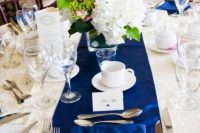 a bold table setting with a navy table runner, a white floral centerpiece with greenery and white candles