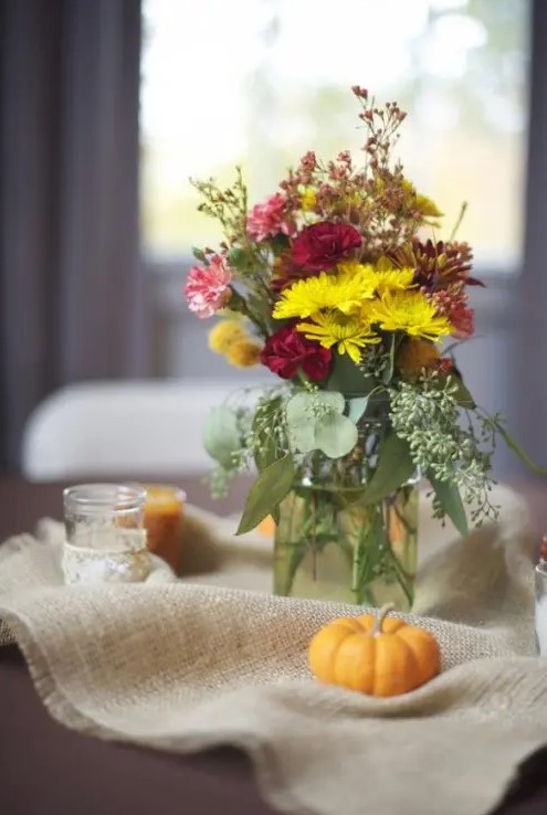 a bright fall wedding centerpiece of burlap, little pumpkins, candles, a jar with bright blooms and greenery will bring a rustic feel
