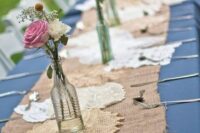 a burlap table runner with doilies, bottles with white and pink blooms and greenery is a cool idea for a rustic wedding
