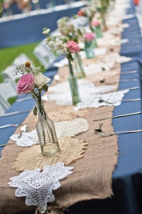 a burlap table runner with doilies, bottles with white and pink blooms and greenery is a cool idea for a rustic wedding